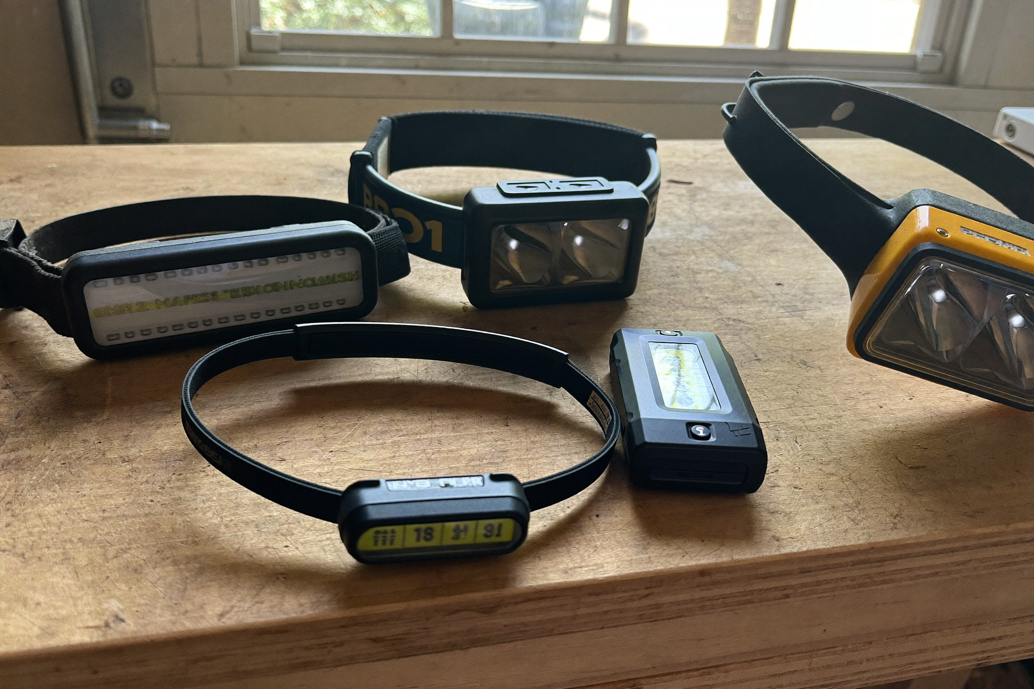 Five different LED headlamps and work lights lined up side by side on a workbench
